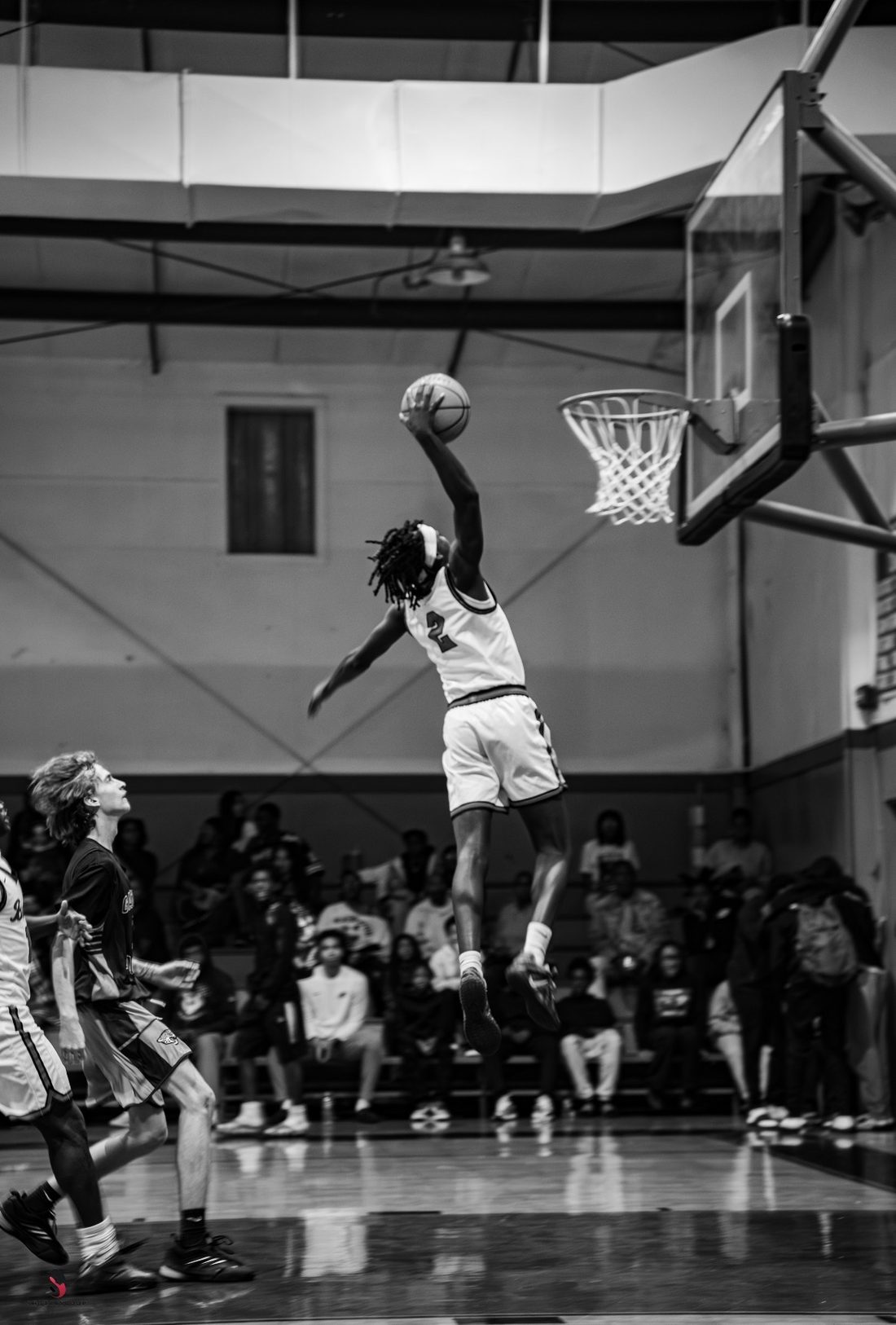 Black and white photo of a basketball player in a white number 2 jersey rising toward the rim for a one-handed dunk in a packed gym