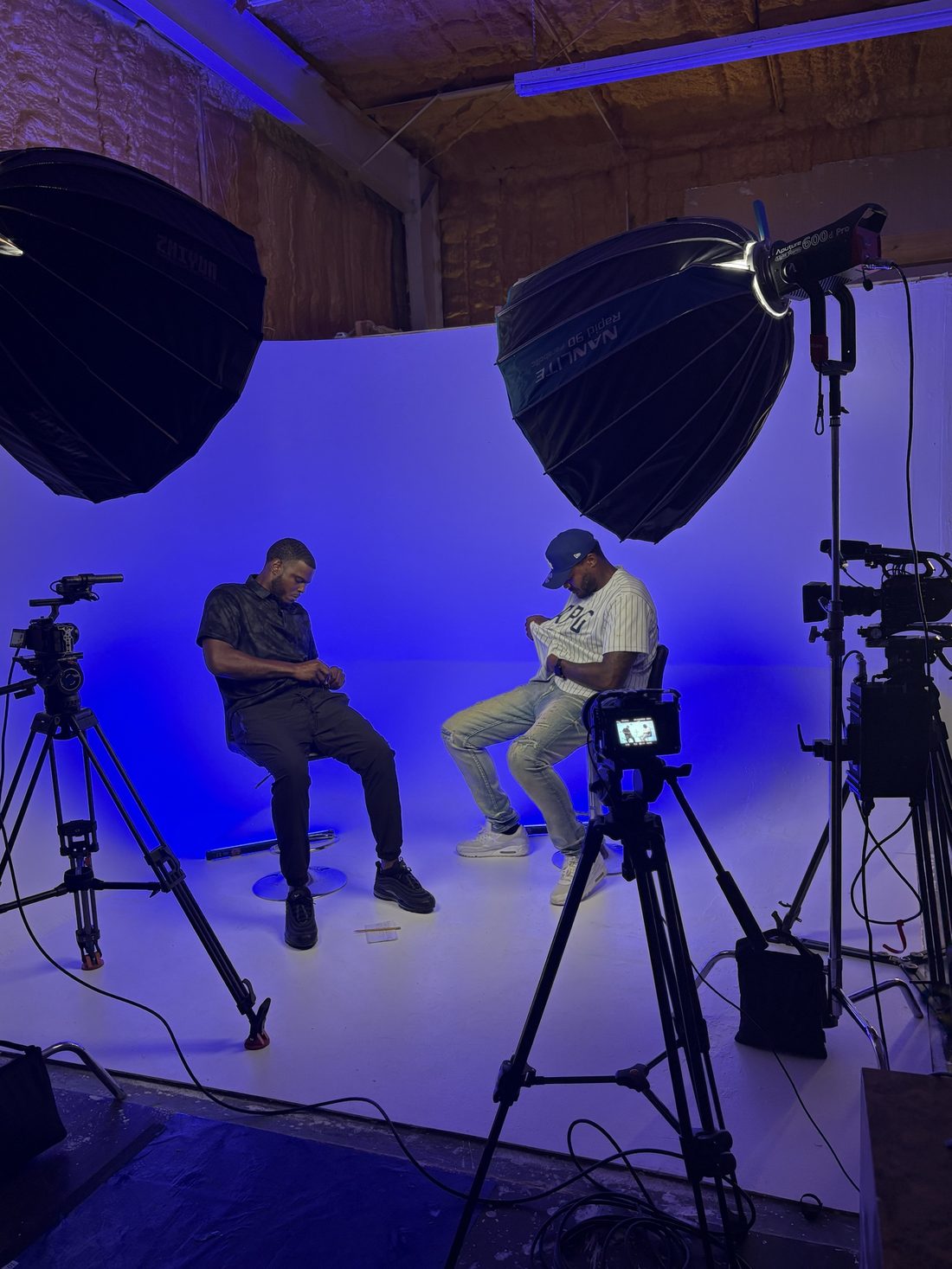 A studio set with two cameras on tripods, large softbox lights and two people seated in front of a blue backdrop