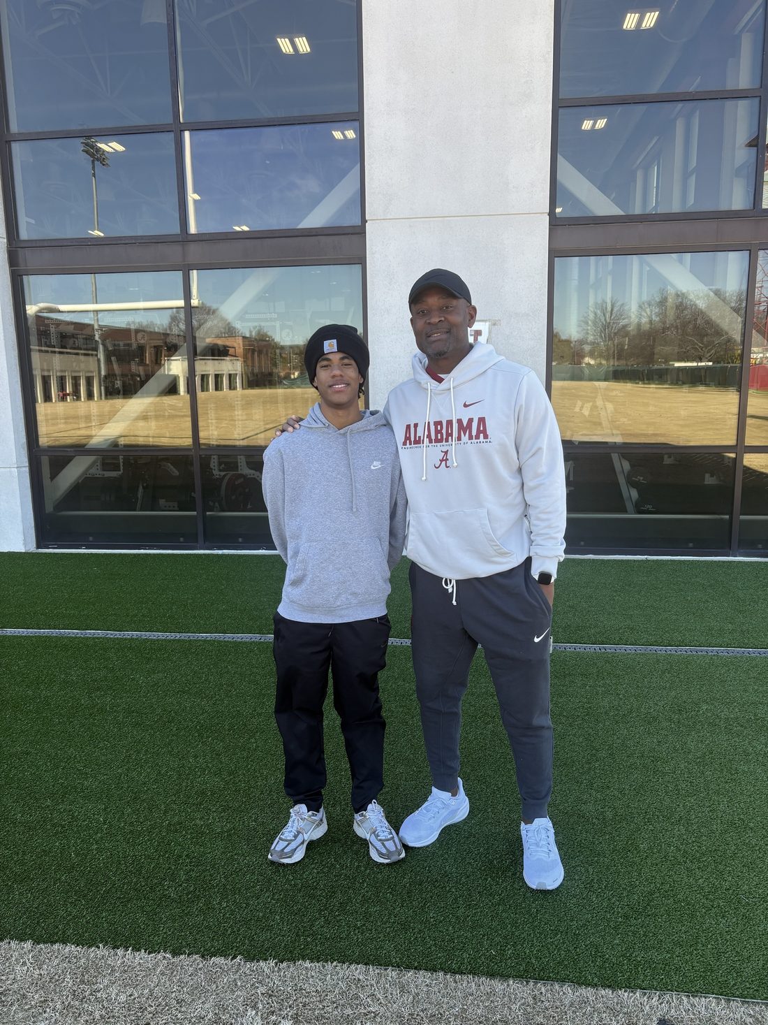 A young athlete and an adult stand together on turf outside a college athletics building